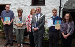 A line of six people stand smiling, three hold blue Cornwall Heritage Awards plaques - from the awards ceremony at Mary Newman's Cottage, Saltash.