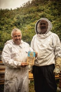 Two men in beekeeper outfits smile and hold a blue Cornwall Heritage Awards trophy.
