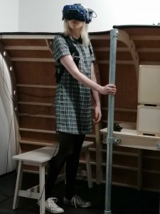 A young blonde woman (Amy) stands in a room and looks around whilst wearing a black VR headset.