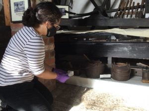 A young woman wearing a mask and gloves kneels before an old hearth, cleaning. 